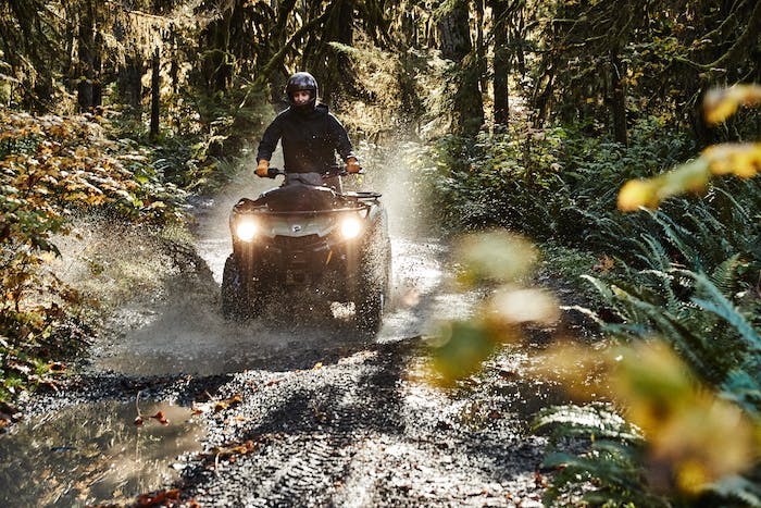 man driving atv splashing through puddle on dirt road in a mossy forest