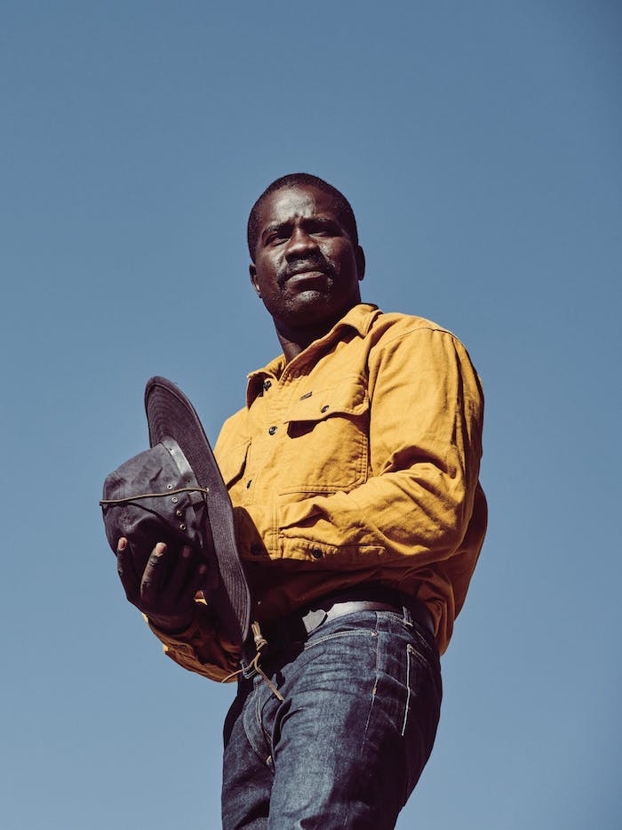 African American man wearing a yellow flannel shirt and holding a oil cloth hat looking off into the distance with a blue sky behind him