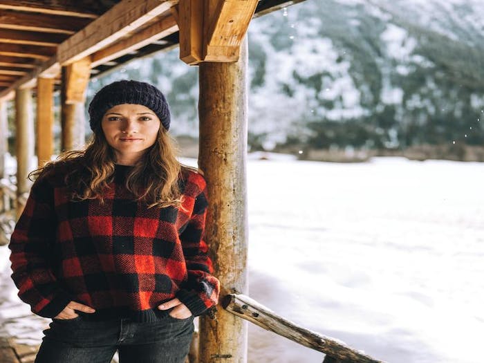 brunette woman standing on the porch of a cabin during a snowy winter wearing a red buffalo plaid sweater and hand knit beanie