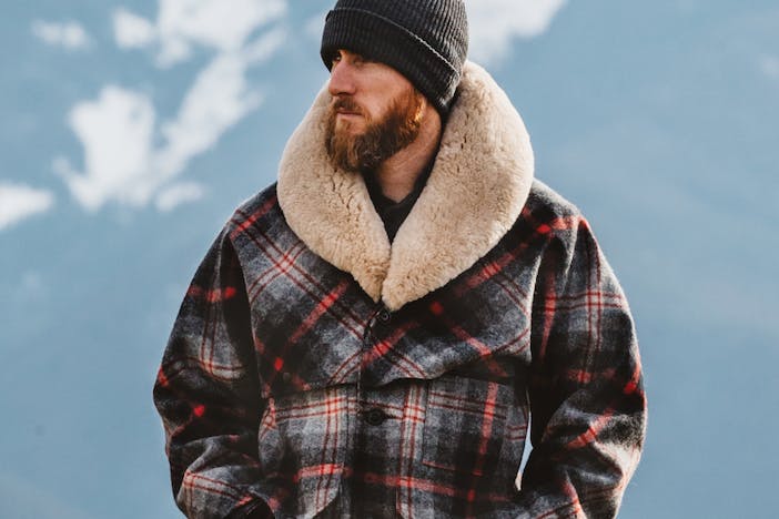 a bearded man wearing a grey beanie and a red, black and white plaid coat with a wool collar looking off to the left