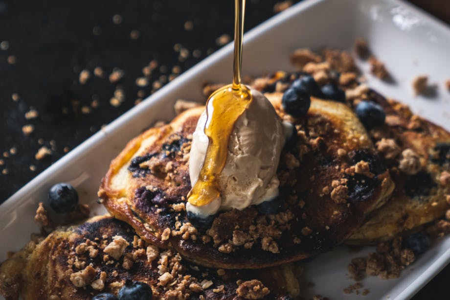 blueberry pancakes with crumbled walnuts on a white plate with syrup drizzling from a tin