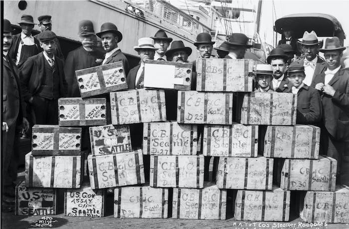 black and white image of people in antiquated clothes and hats standing behind a large stack of bound wooden boxes reading 