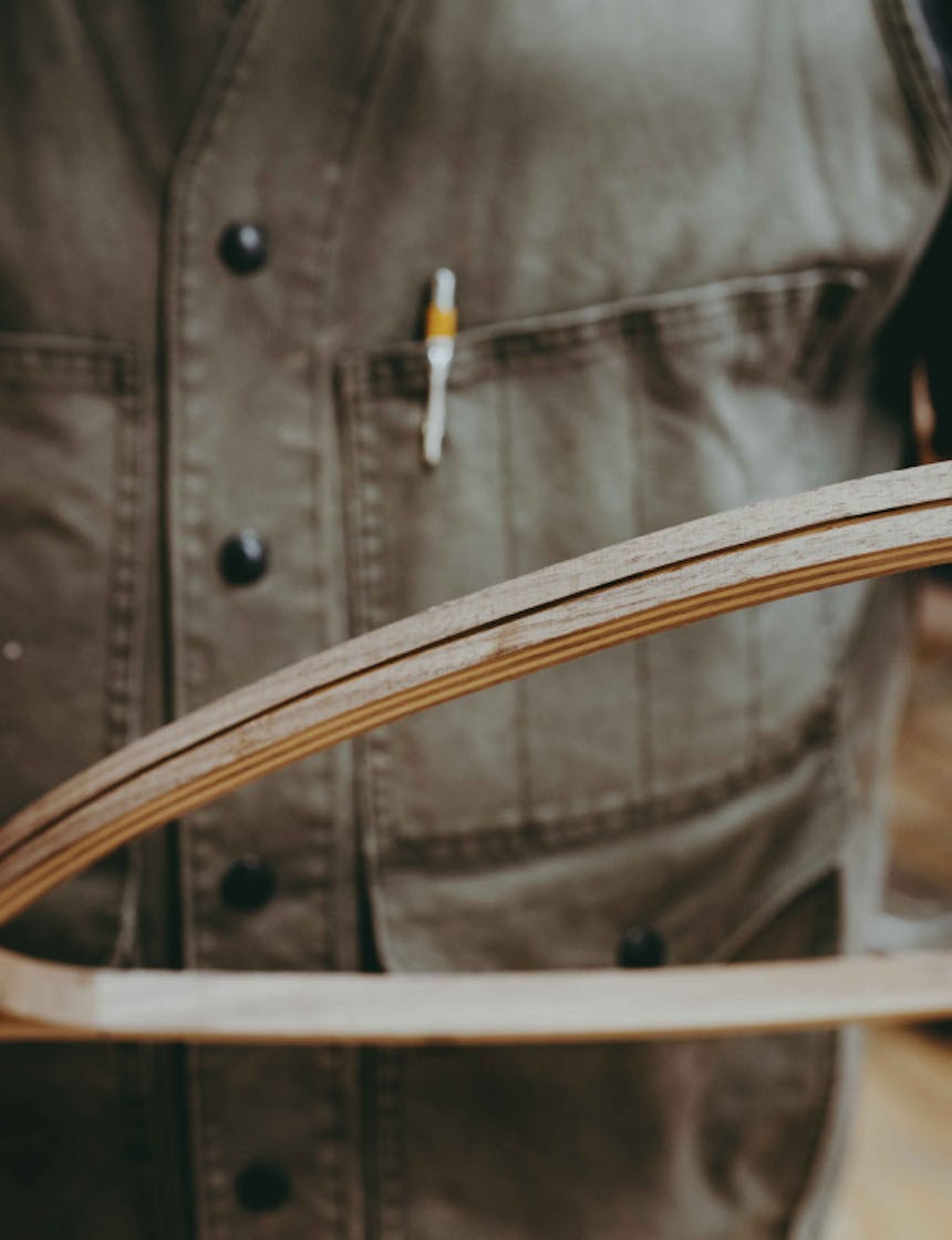 a close up of the woodworker wearing a green vest with the yellow mechanical pencil in his chest pocket while he holds the wooden net close to the camera