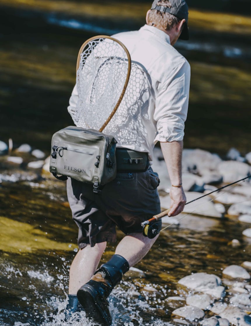 view from behind a white man wearing a white long sleeve button up with the sleeves rolled, dark grey shorts, a green dry waist pack with a wooden fishing net tucked in while he holds a fly fishing rod walking into a river