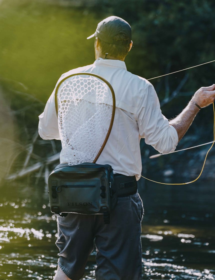 view from behind a white man wearing a white long sleeve button up with the sleeves rolled, dark grey shorts, a green dry waist pack with a wooden fishing net tucked in while he casts a fly fishing rod standing in a river