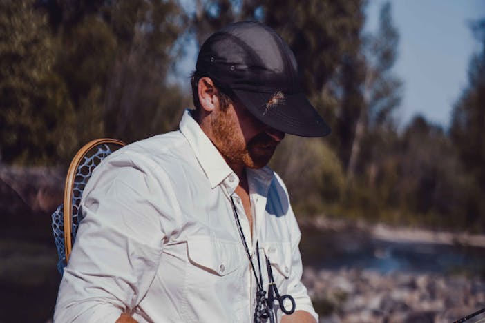 a side view of a white fisherman wearing a white button-up, black duck billed brim with a fly stuck in it and the landing net tucked into his waist pack