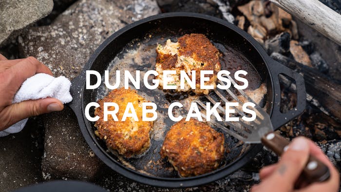 hand holding a cast iron skillet with three dungeness crab cakes over the coals of a fire central text reads 