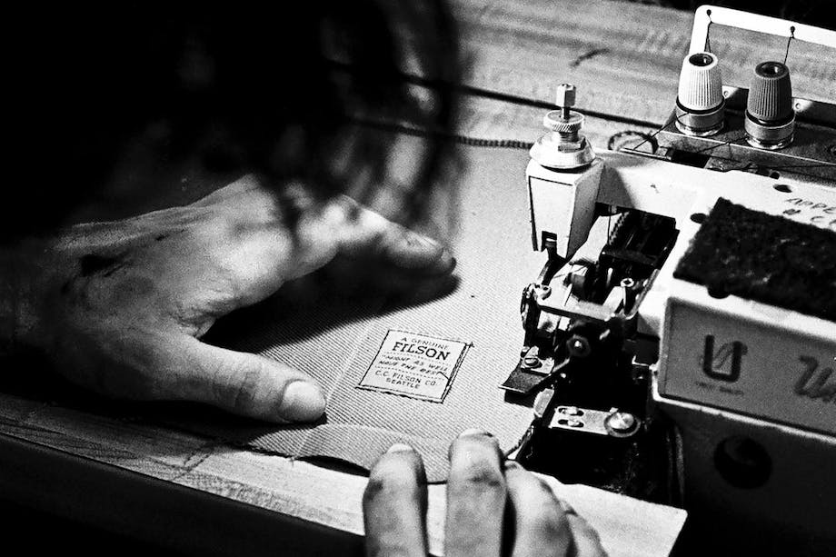 an archival black and white image from behind the sewer working on sewing two pieces of fabric together on an old sewing machine with the original Filson logo already sewn on the bottom right corner.