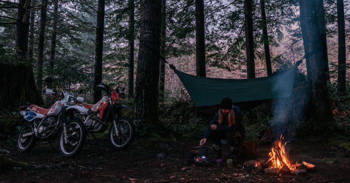 person squats next to a fire in a campsite with two dirtbikes and pinetrees