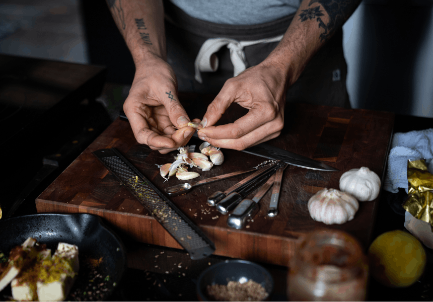 hands removing garlic peels over a wooden cutting board with measuring tools and a microplane