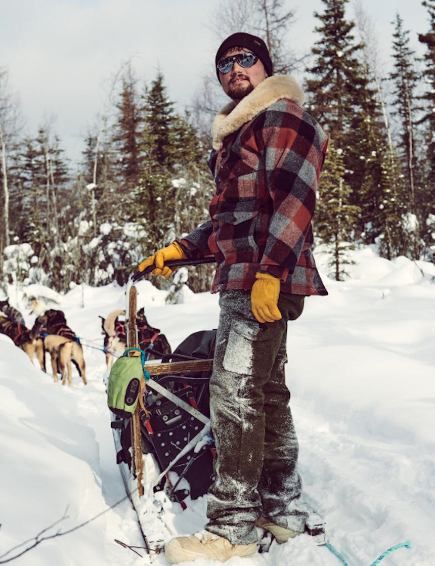 a man standing on the back of a dog sled wearing a black, grey and red plaid wool coat and green wool pants while the team waits for a command