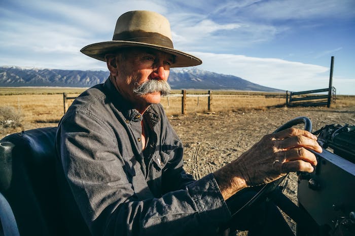 old farmer with tan flat billed hat in grassland on farm vehicle and mountain backdrop