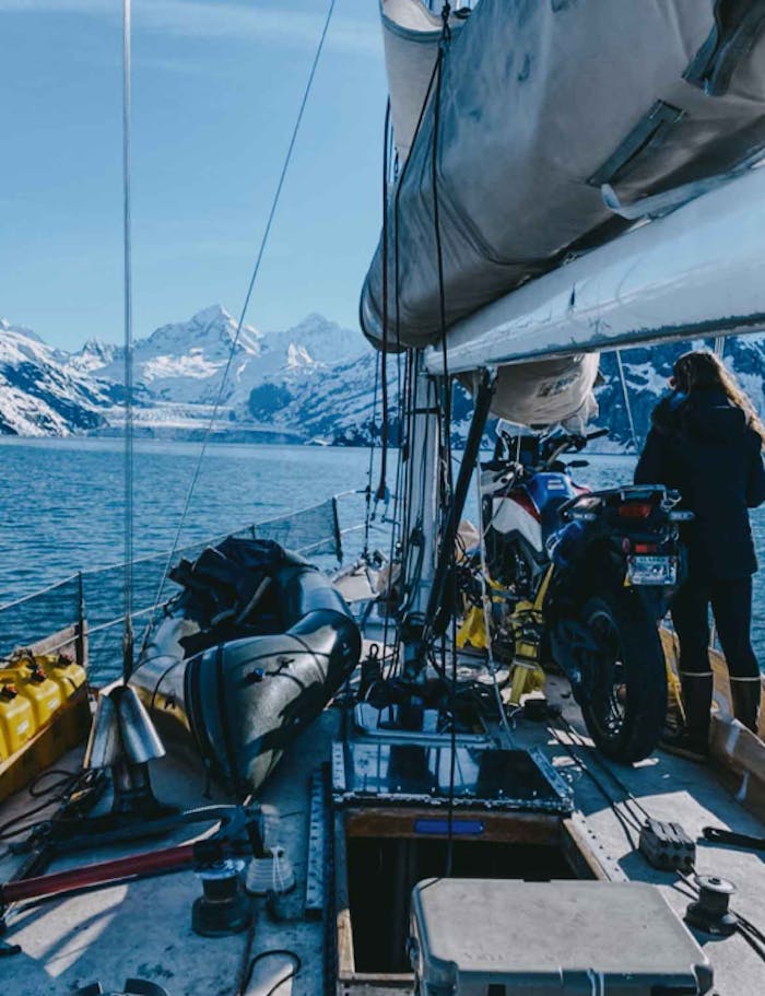 view from the deck of a working sailboat that has gear and a motorcycle strapped to the deck while a woman looks out to the snowy mountains