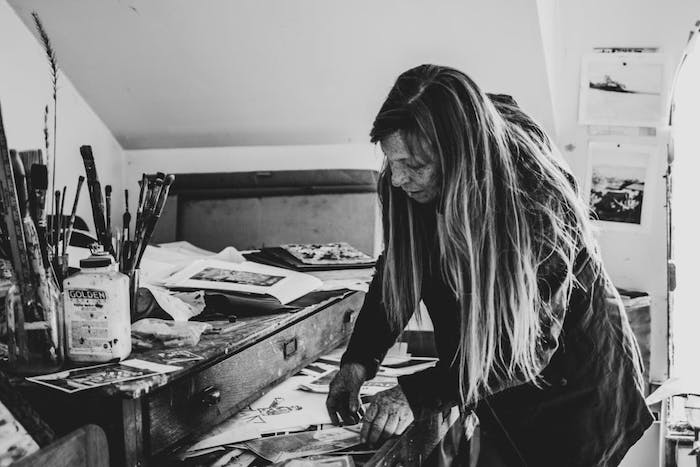 black and white image of woman in art studio with assorted paint brushes and reference images strewn about and tacked to wall