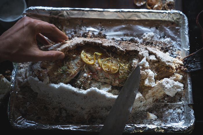 hand removing salt crust from a baked fish with lemon and herbs exposed on top of the fish skin