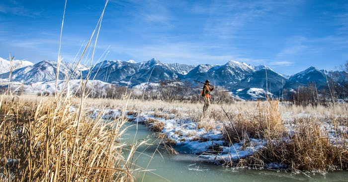 Hunter in high visibility vest with shotgun walking through snowy grassland with river with mountain backdrop